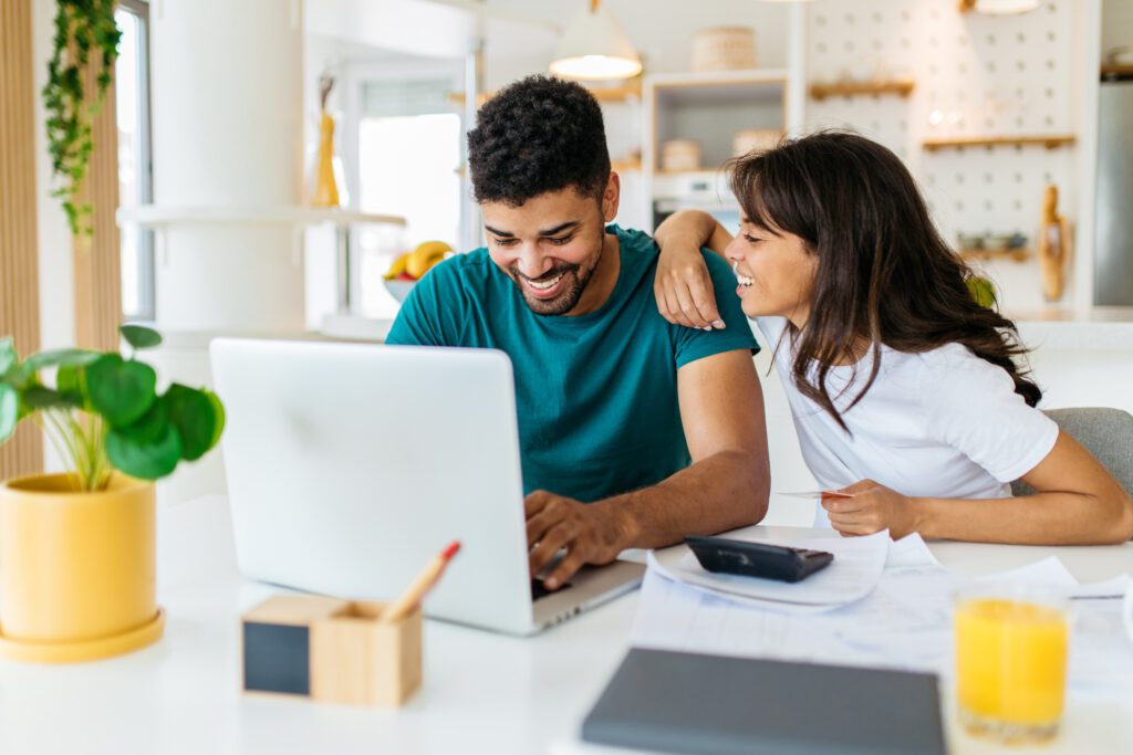 A young responsible African American couple sits at home and calculating monthly income. The man is looking at the laptop while the woman explaining to him how to save some money.