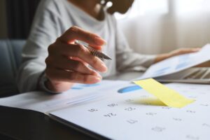 Asian woman is writing on yellow paper notes and put a warning on calendar. The other hand hold the report graph and has laptop on the front. Maybe preparing data for marketing report, work from home