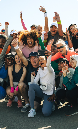A diverse, energetic group of runners poses for a joyful selfie, smiling and raising their arms in celebration against a bright outdoor backdrop.