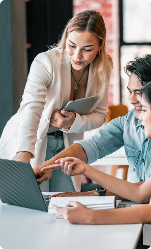 A woman in a white blazer, holding a tablet, leans over a desk to assist two smiling colleagues pointing at a laptop screen, suggesting teamwork.