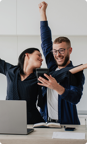 A joyful man and woman celebrate in an office, raising their arms triumphantly while looking at a tablet. A laptop and notebook are on the desk.