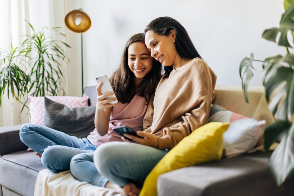 A mother and daughter sit on a cozy couch, smiling at a smartphone. The room is bright with plants and colorful cushions, creating a warm, joyful atmosphere.