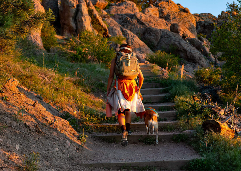 Woman in hiking gear and bookbag, walking dog, climbing up stairs on side of mountain